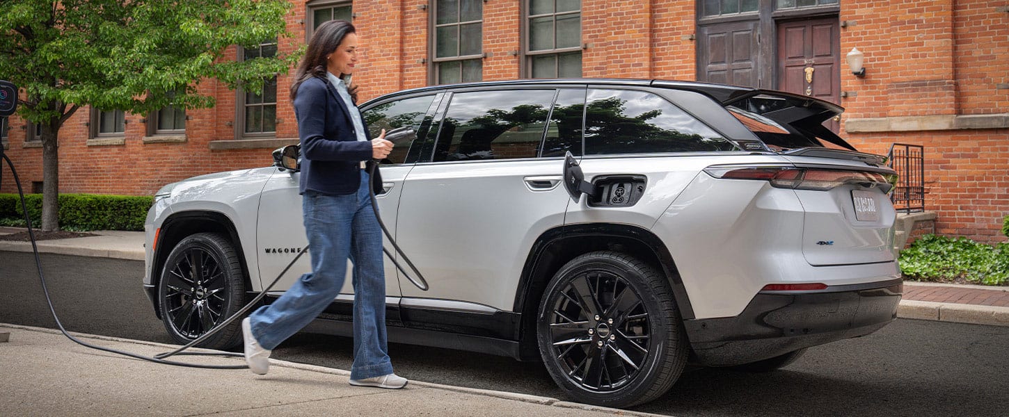 A woman walking toward a silver 2025 Jeep Wagoneer S Launch Edition with its charging port door open and a charging cord in her hand.