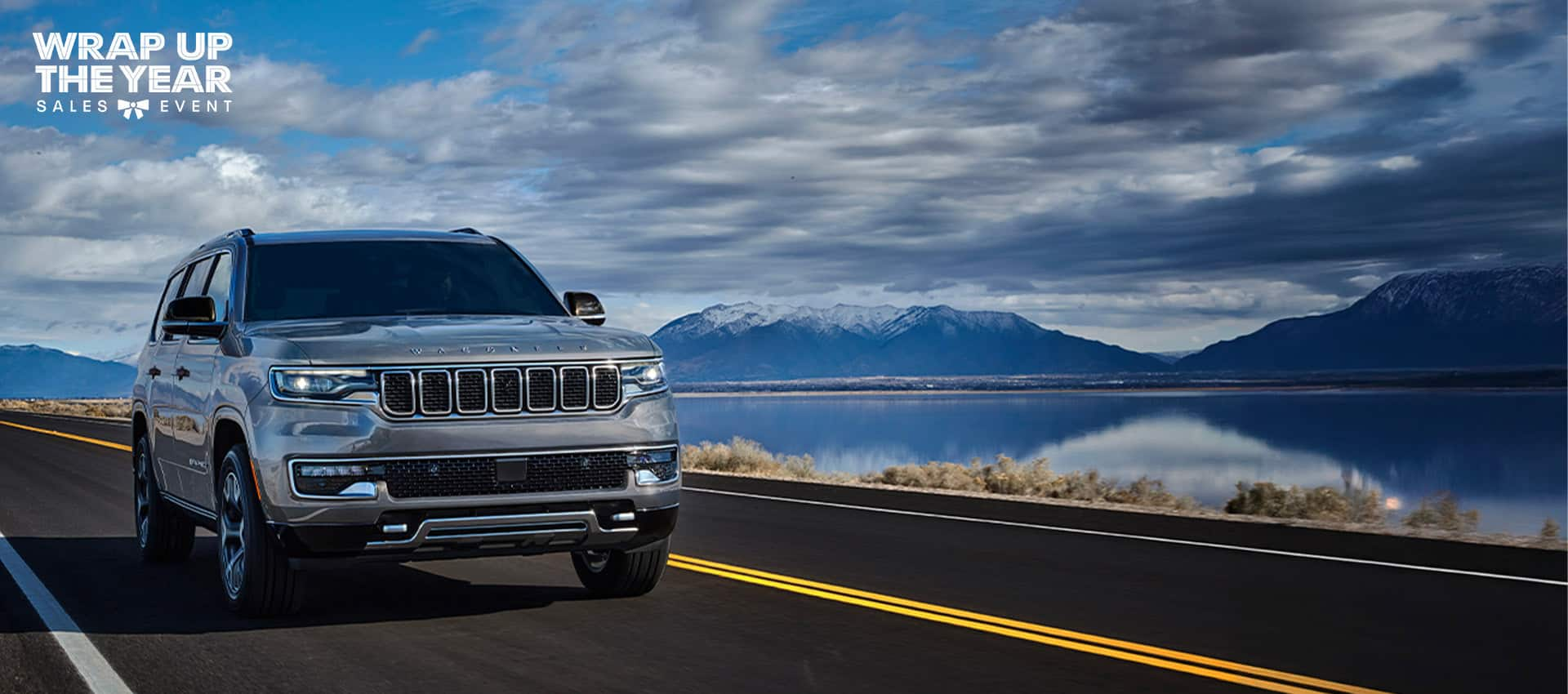 A silver 2025 Jeep Wagoneer Series III traveling on a highway beside a lake, with mountains in the background. Wrap Up The Year Sales Event logo.