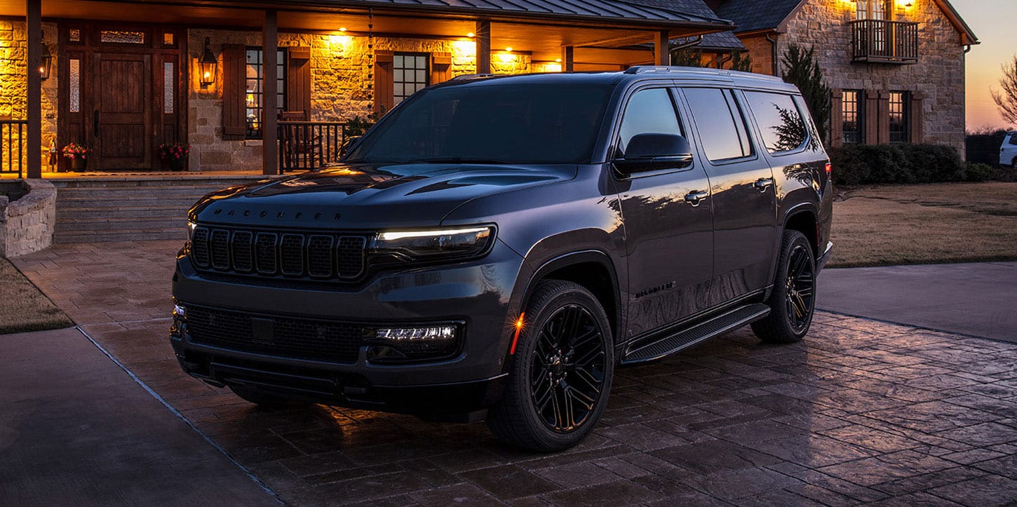 A driver-side front angle of a black 2025 Jeep Wagoneer Carbide with its Daytime Running Lamps on, parked in the courtyard of a large, upscale lodge at sunset.