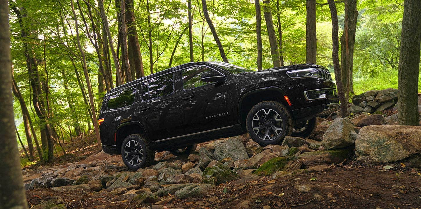 A passenger-side profile of a black 2025 Jeep Wagoneer crawling up a rocky hill off-road, in the woods.