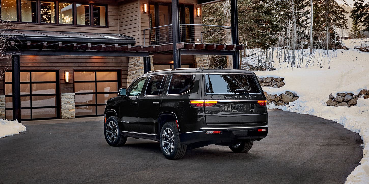 A driver-side rear angle of a black 2025 Jeep Wagoneer Series III parked in the driveway of a luxurious mountain chalet with snow-covered woods in the background.