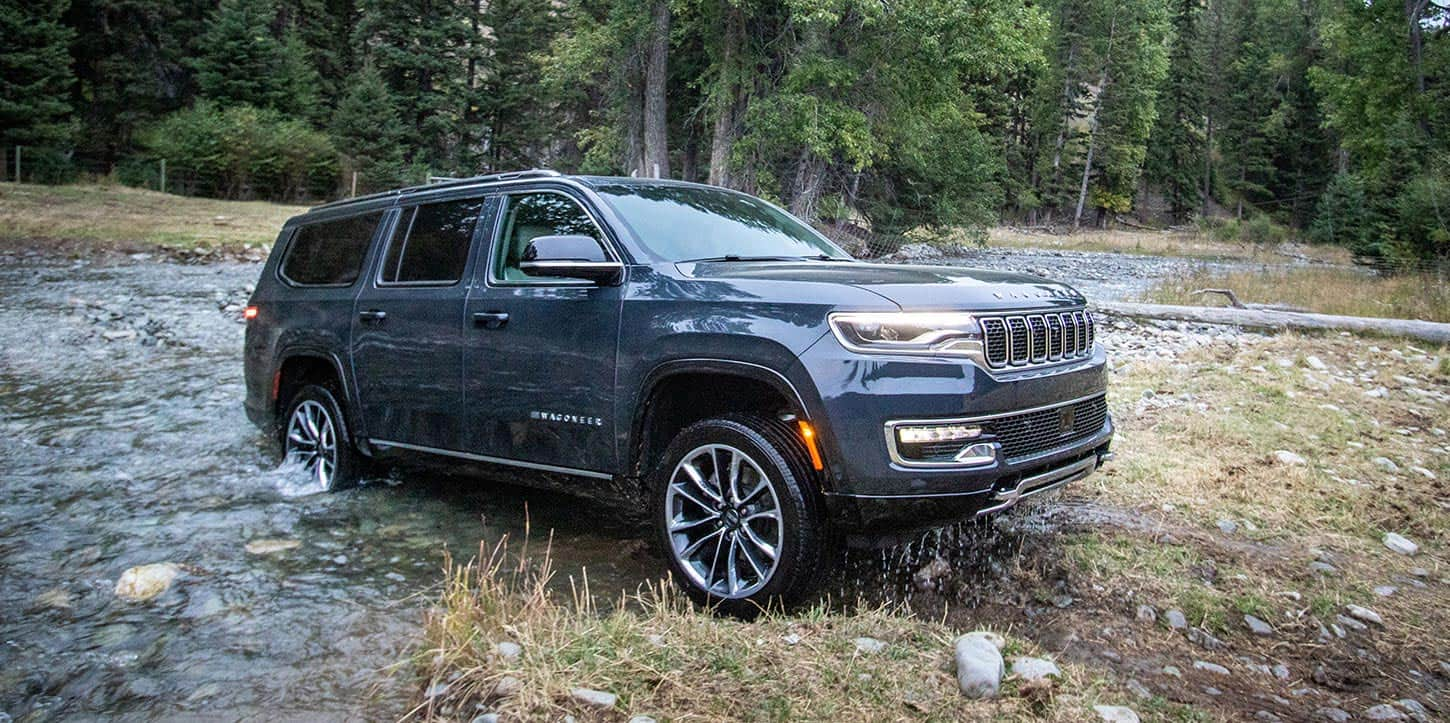 A passenger-side angle of a blue 2025 Jeep Wagoneer exiting a stream on a trail off-road in the woods.