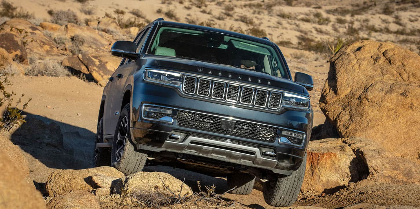 A front end angle of a gray 2025 Jeep Wagoneer Series III ascending a rocky hill with the tires crawling over an uneven surface of different size boulders.