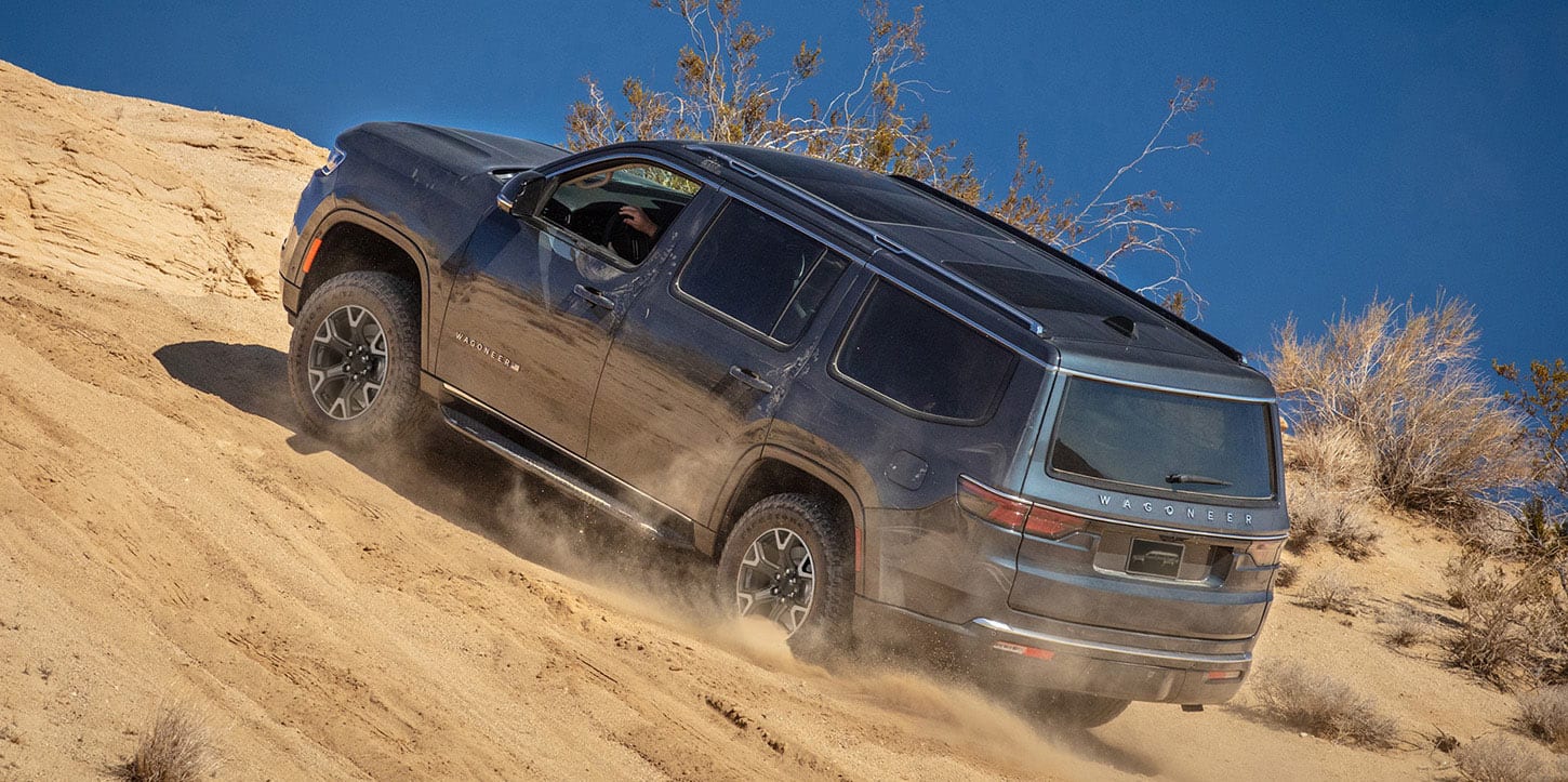 A driver-side profile of a gray 2025 Jeep Wagoneer crawling up a very steep, sandy hill off-road, in the desert.