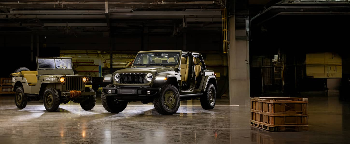 A historic military Jeep vehicle and a 2025 Jeep Wrangler Willys '41 4xe Special Edition with its doors and roof removed, parked perpendicular to each other in a darkened studio.