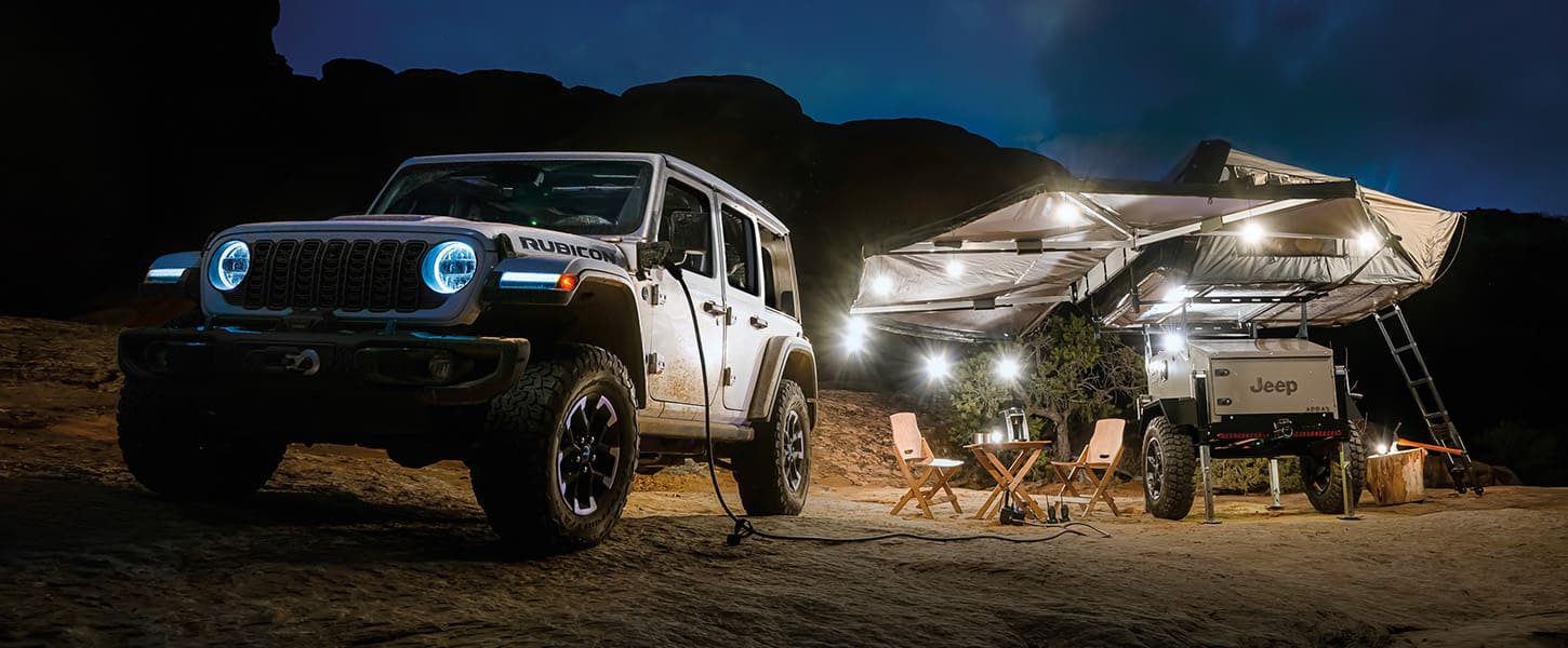 Un Jeep Wrangler Rubicon X 4xe 2025 estacionado fuera de la carretera, en el desierto, por la noche, con un extremo de un cable de alimentación en su caja de alimentación y al lado de una casa rodante.