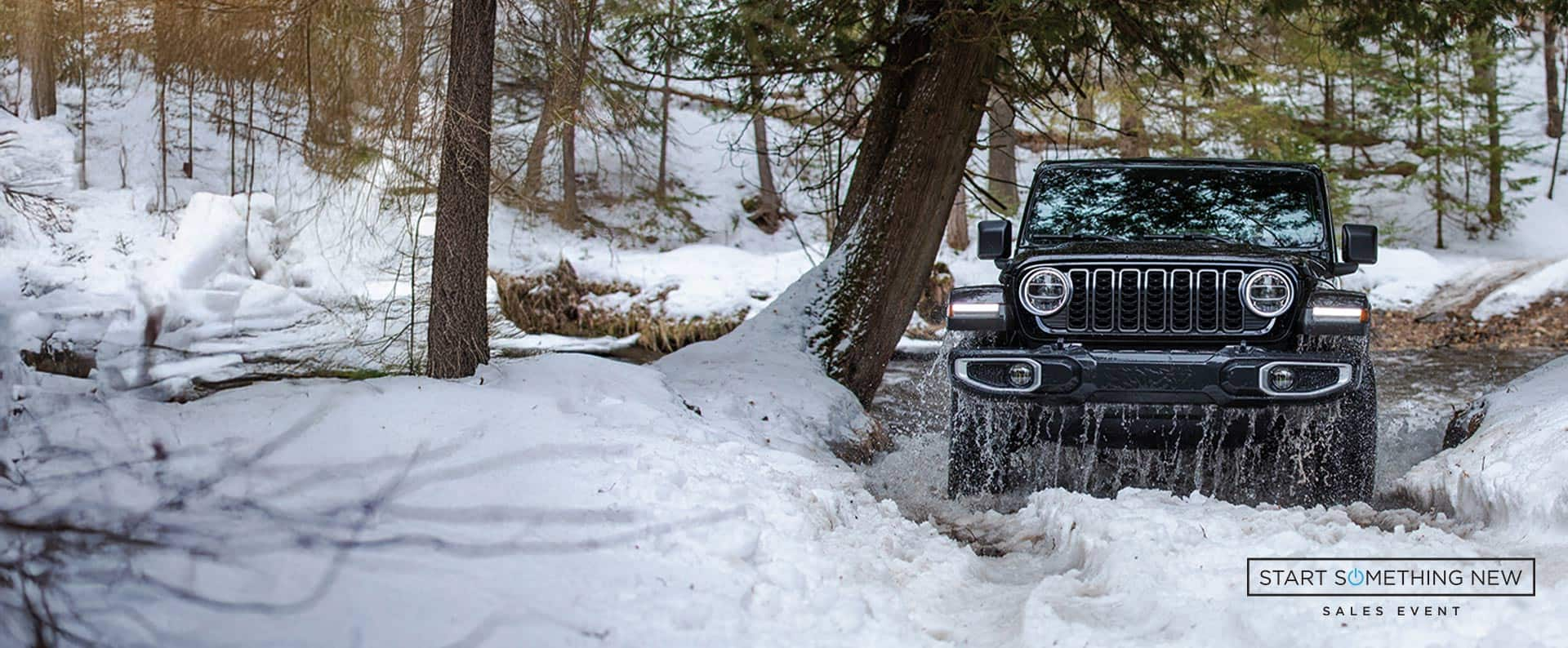 A front angle of a black 2025 Jeep Wrangler Sahara exiting an icy stream off-road, in a snow-covered forest. Start Something New Sales Event logo.