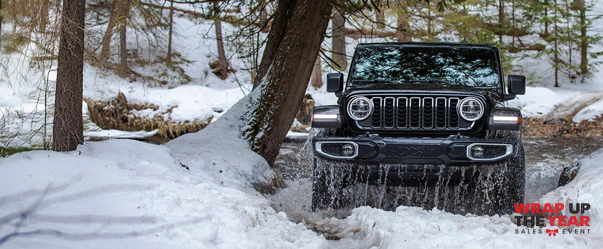 A front angle of a black 2025 Jeep Wrangler Sahara exiting an icy stream off-road, in a snow-covered forest. Wrap Up The Year Sales Event logo.