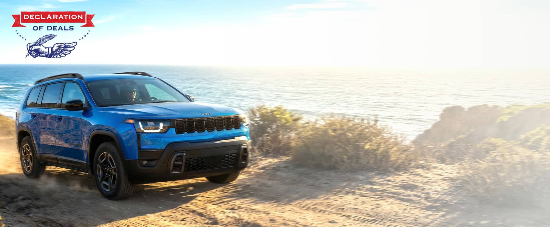 A passenger-side front angle of a blue 2026 Jeep Cherokee Laredo traveling on a beach beside the ocean. Declaration of Deals.
