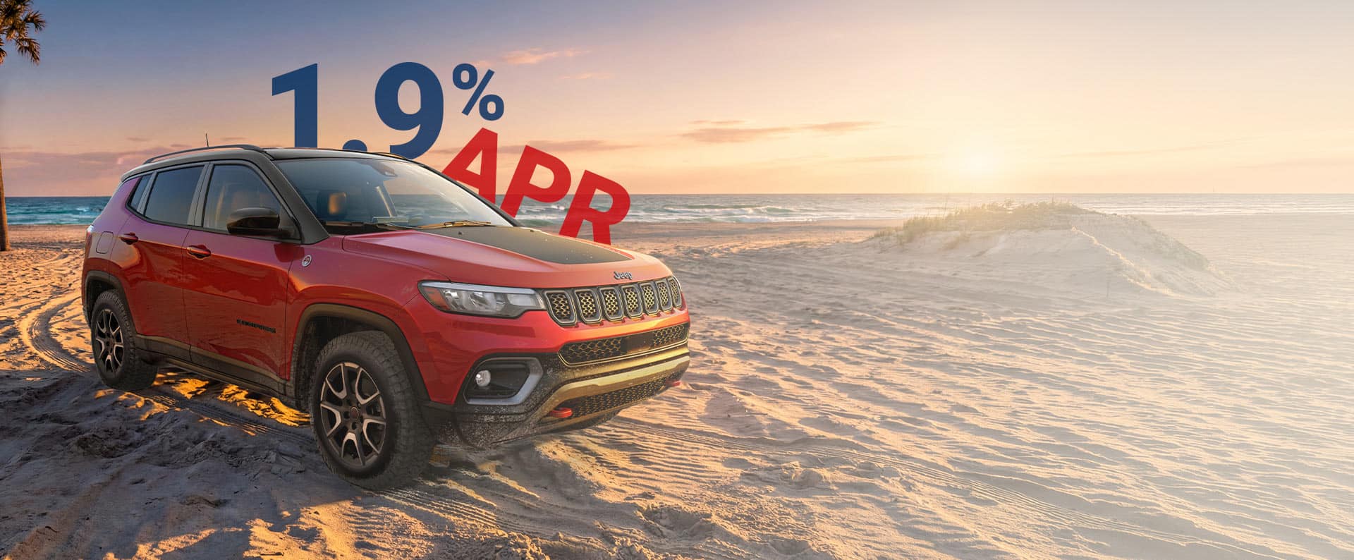 A red 2026 Jeep Compass Trailhawk with black hood insert, parked on a sandy beach at sunset. 1.9% APR.
