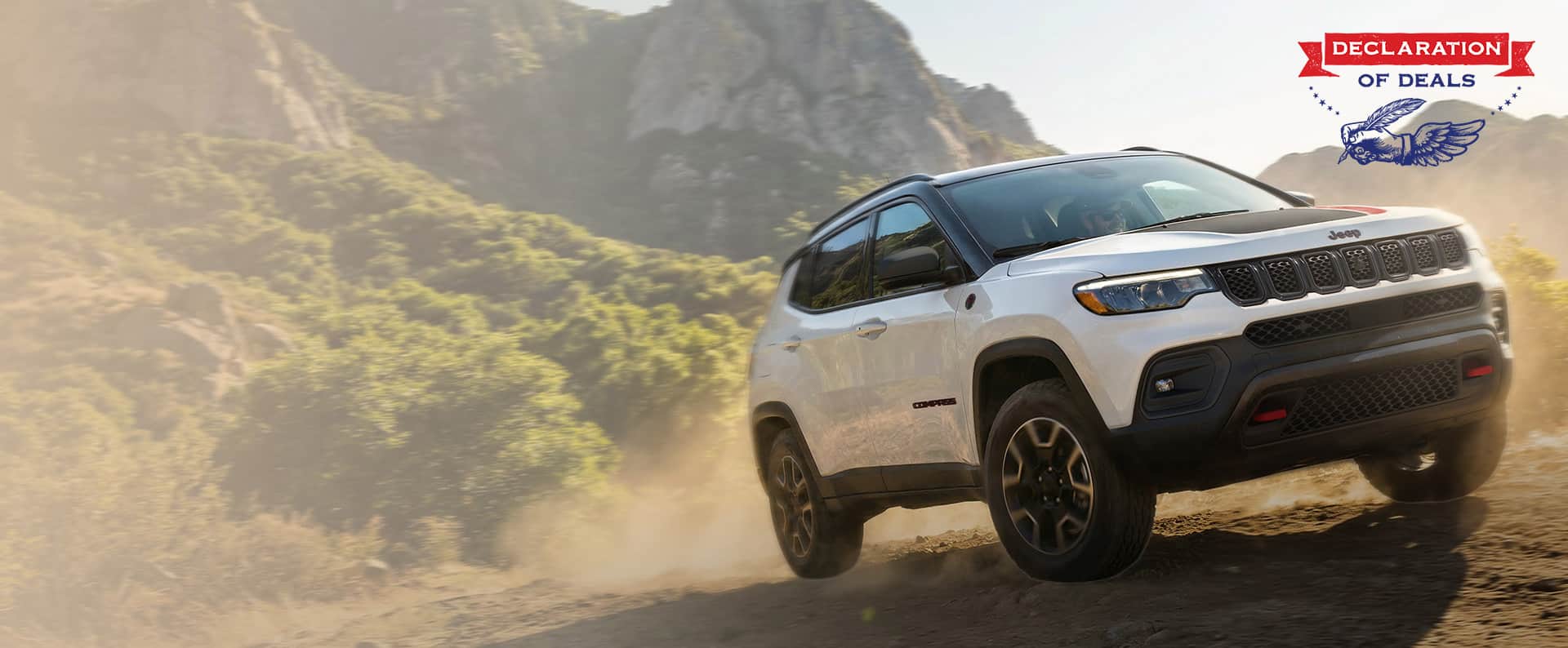 A passenger-side front angle of a white 2026 Jeep Compass Trailhawk with black hood insert ascending a trail off-road. Declaration of Deals.