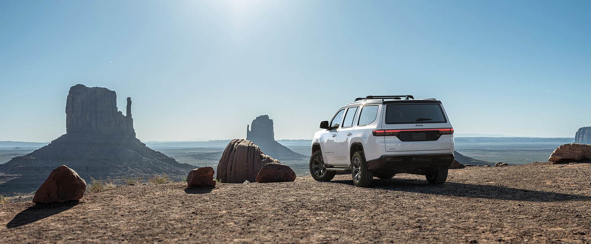 A driver-side rear angle of a white 2026 Jeep Grand Wagoneer Upland parked on a sandy clearing with large rock formations in the background.