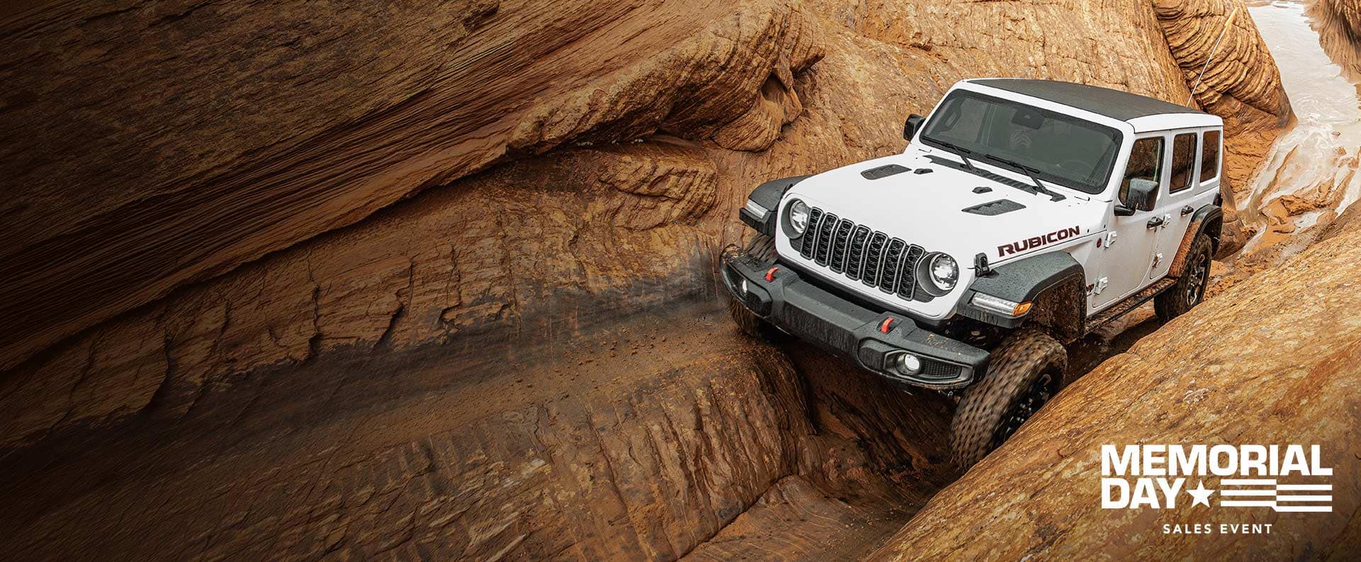 A front angle of a white 2026 Jeep Wrangler Rubicon crawling through an incredibly rugged, narrow stone trail. Memorial Day Sales Event.