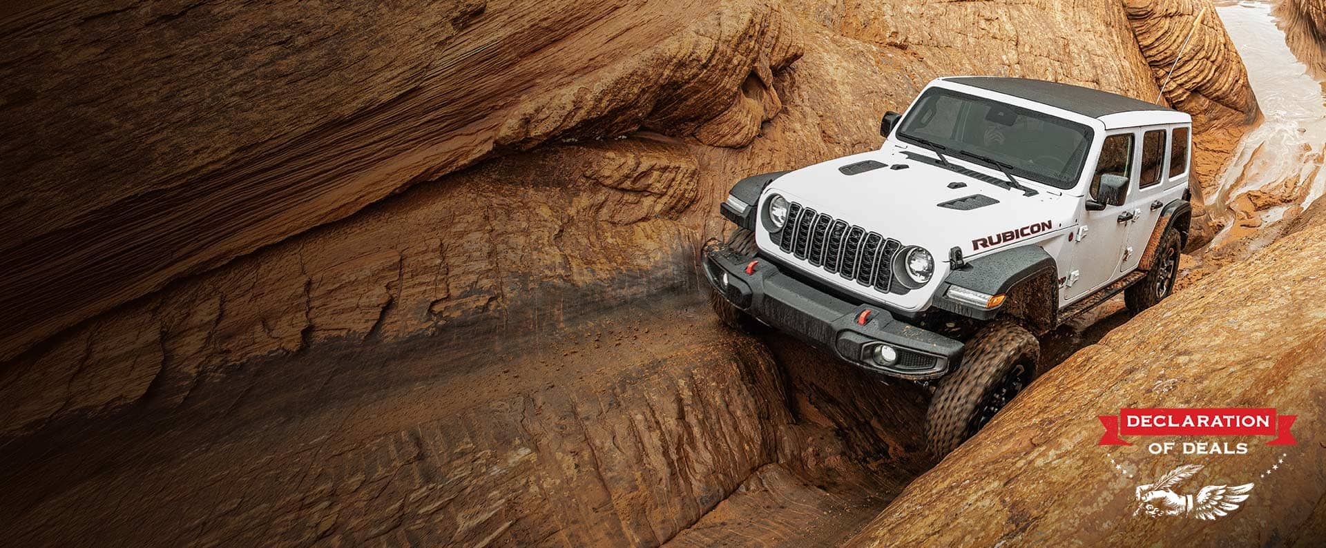 A front angle of a white 2026 Jeep Wrangler Rubicon crawling through an incredibly rugged, narrow stone trail. Declaration of Deals.