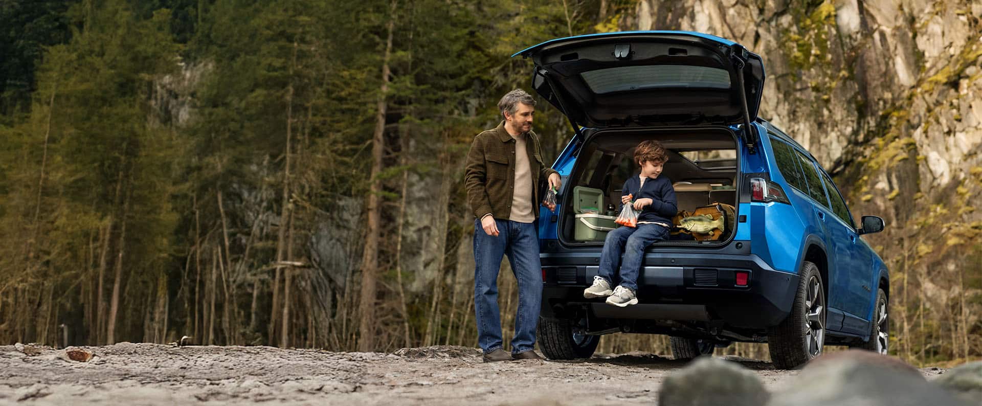 The rear of a blue 2026 Jeep Cherokee Overland parked on a clearing with its liftgate open and a young boy sitting inside with a Billy Bass wall mount beside him along with fishing gear. The father is standing beside the open liftgate. Both are looking at Billy Bass. A wooded swamp is in the background.