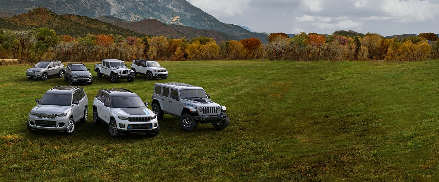 A lineup of seven 2022 Jeep Brand vehicles parked on a grassy plain with a forest wrapped in autumn leaves and mountains rising in the distance.