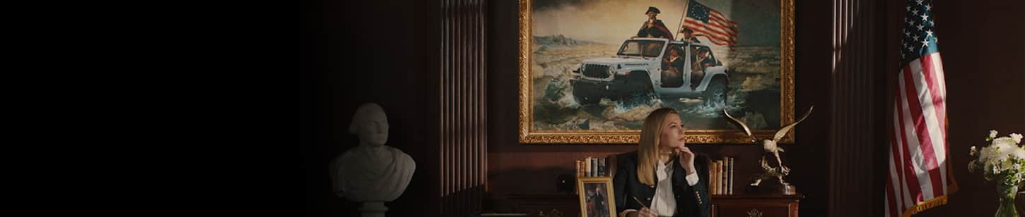A woman at a desk in a government office with a painting behind her of George Washington with three soldiers crossing a shallow part of the Delaware River in a white Wrangler with its doors and top removed.