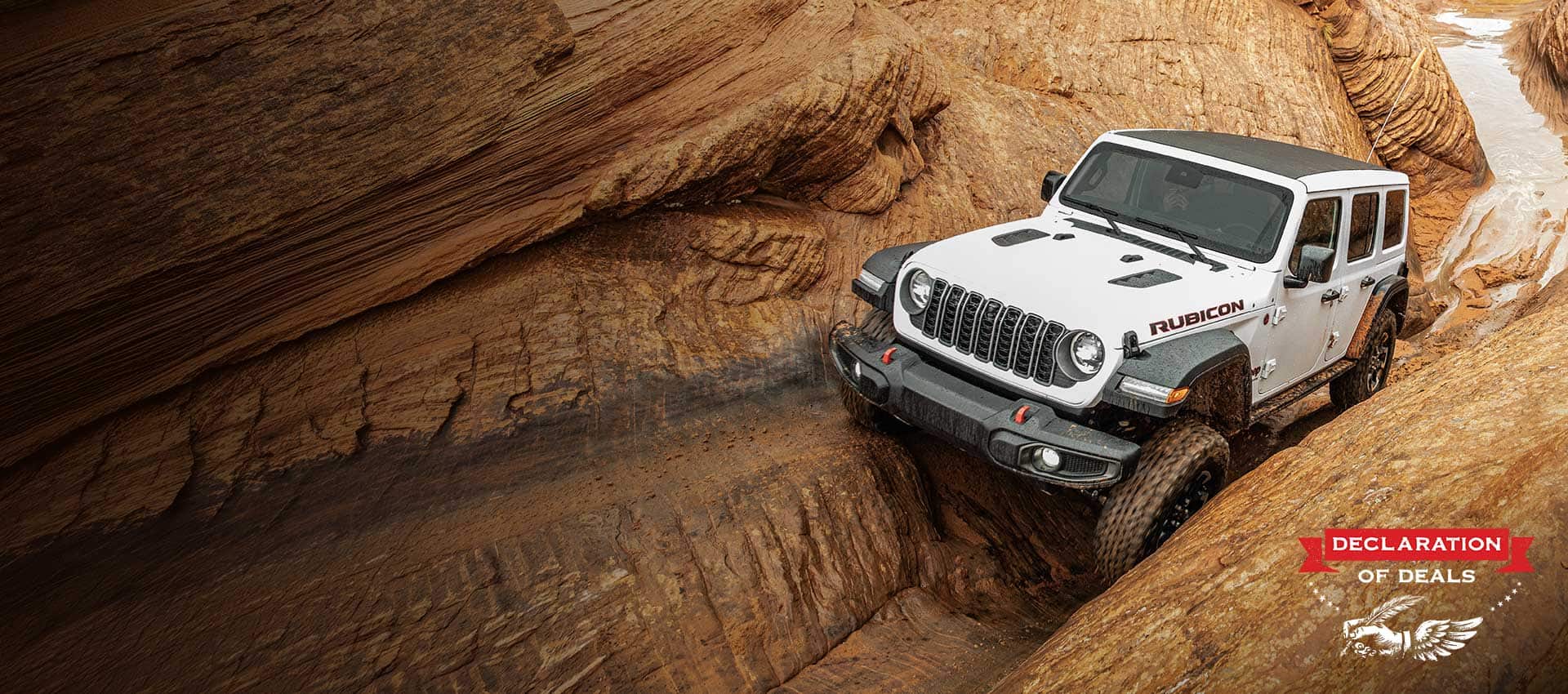 A front angle of a white 2026 Jeep Wrangler Rubicon crawling through an incredibly rugged, narrow stone trail. Declaration of Deals.