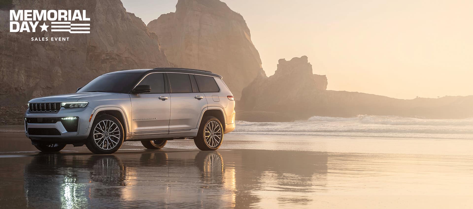 A silver 2026 Jeep Grand Cherokee Summit, parked on a wet beach with rock formations in the background. The Memorial Day Sales Event.