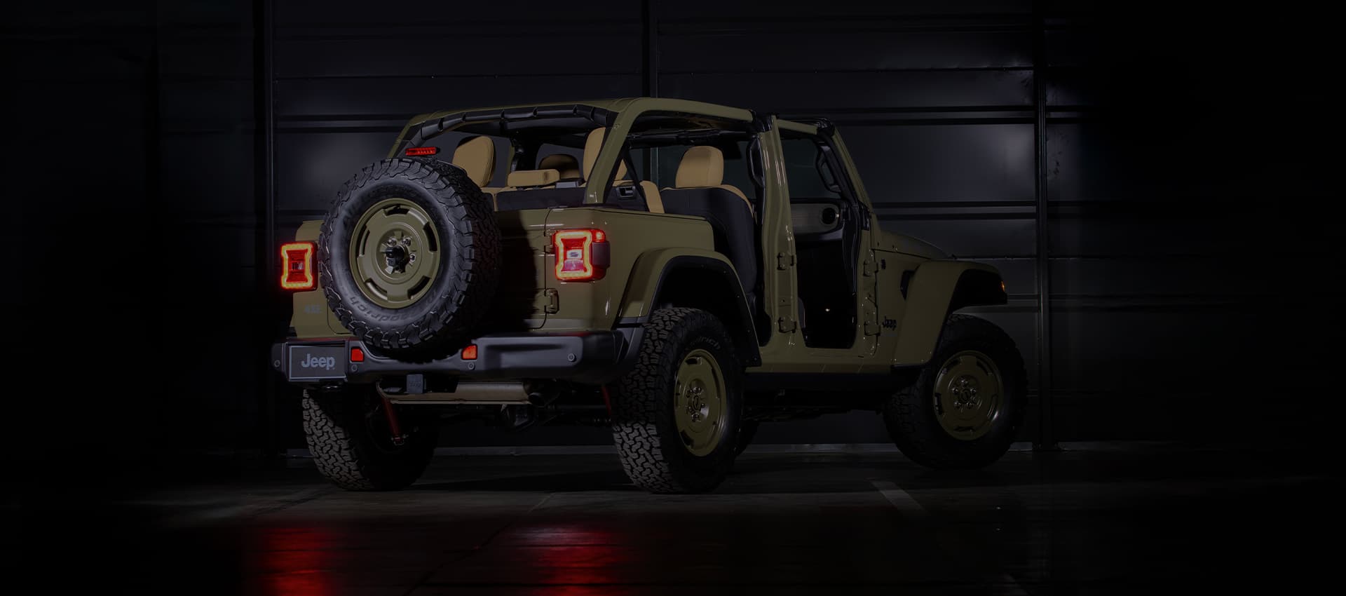 A rear angle of an olive drab 2025 Jeep Wrangler Willys '41 4xe Special Edition with its doors and roof removed, in a darkened studio.