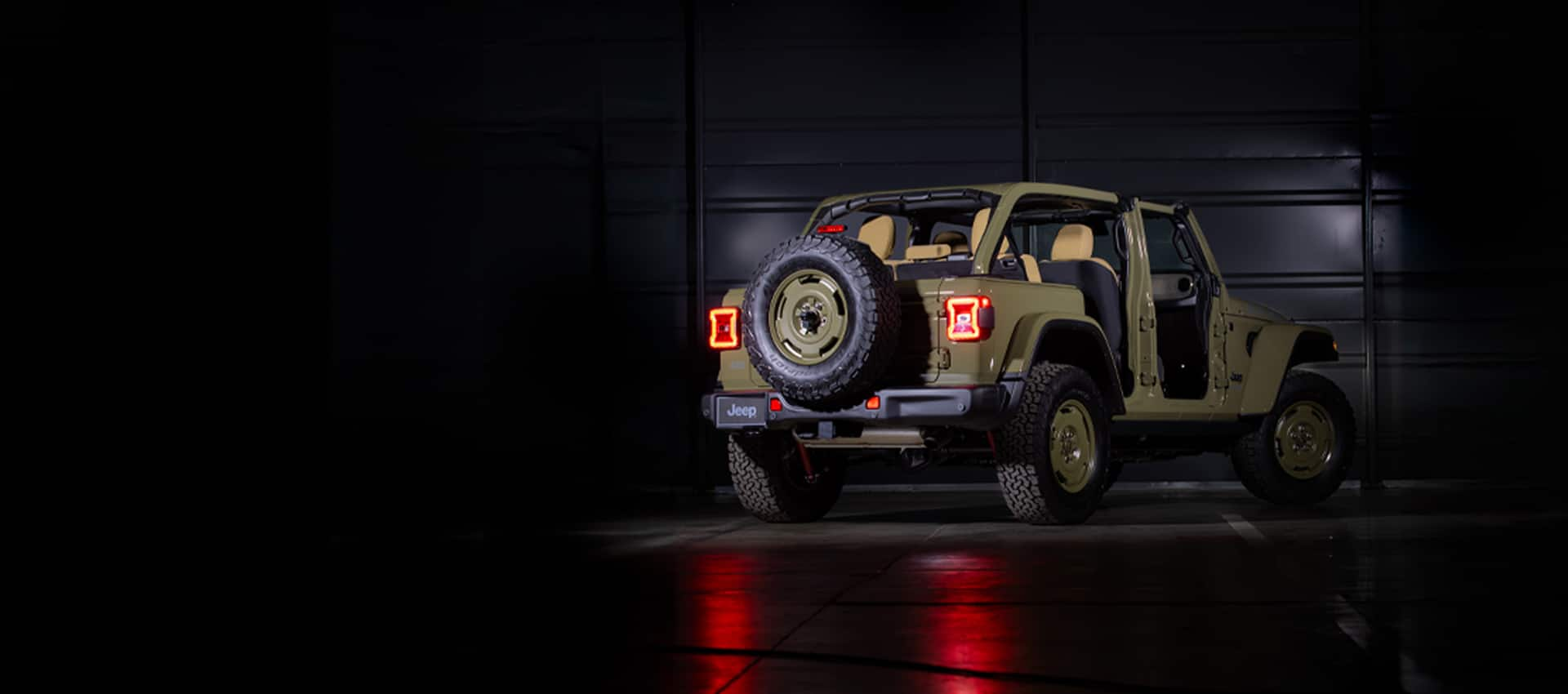 A rear angle of an olive drab 2025 Jeep Wrangler Willys '41 4xe Special Edition with its doors and roof removed, in a darkened studio.