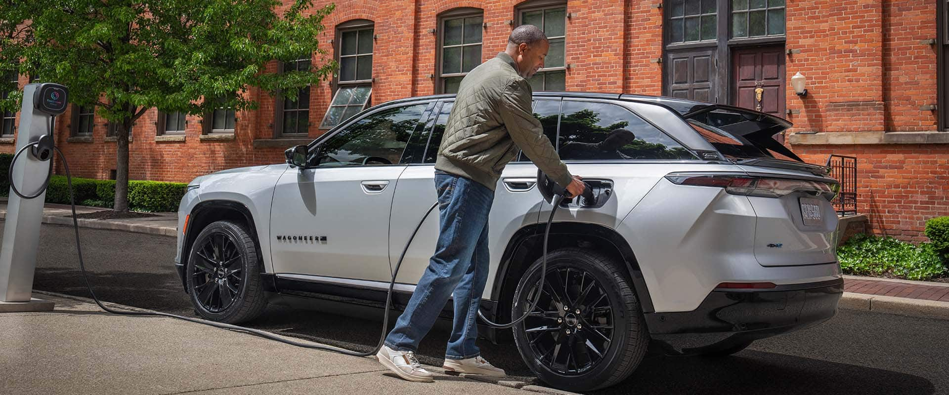 A silver 2024 Jeep Wagoneer S Launch Edition parked beside a brick building as a man plugs the charging cord from a public charging station into the vehicle's charging port. 