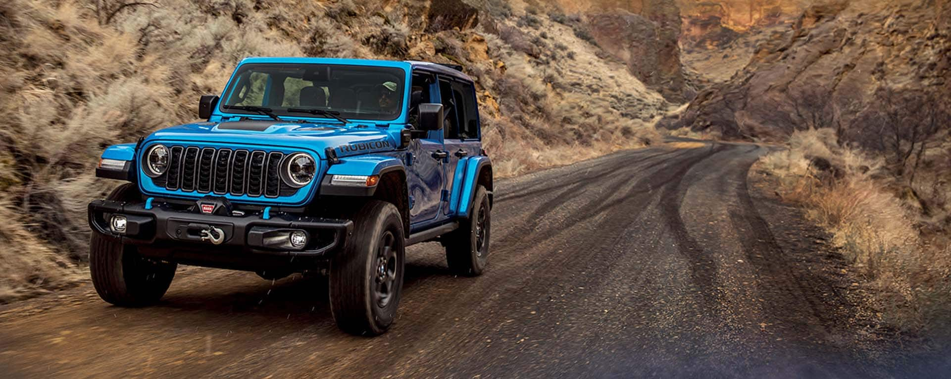 A blue 2024 Jeep Wrangler Rubicon 4xe being driven on a trail cut through the mountains.