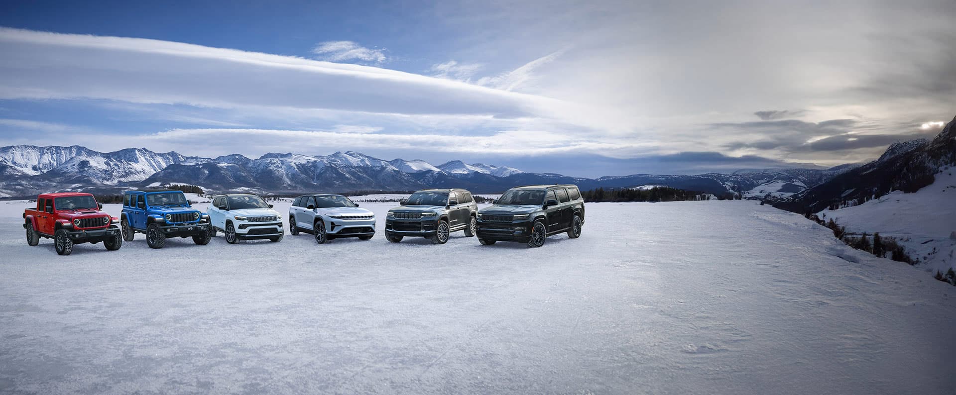 A lineup of six Jeep Brand vehicles parked in a semicircle on a snow-covered clearing in the mountains. From left to right: a red 2025 Jeep Gladiator Mojave X, a blue 2025 Jeep Wrangler Rubicon X 4xe, a white 2025 Jeep Compass Limited, a grey 2025 Wagoneer S, a black 2025 Grand Cherokee Summit Reserve and a black 2025 Jeep Grand Wagoneer Obsidian.