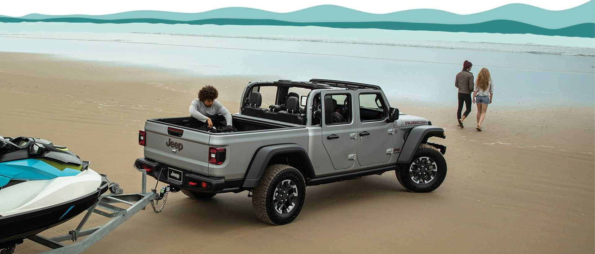 A silver 2024 Jeep Gladiator Rubicon pickup truck with its roof removed, parked on a beach, towing a boat, with a man pulling something out of the truck bed and a couple walking toward the water.