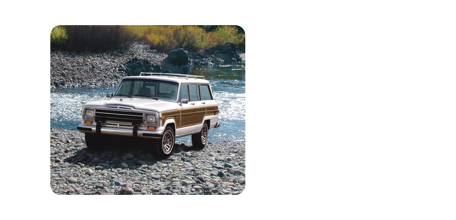 A white Grand Wagoneer with wood side panels, exiting a stream onto a rocky beach.