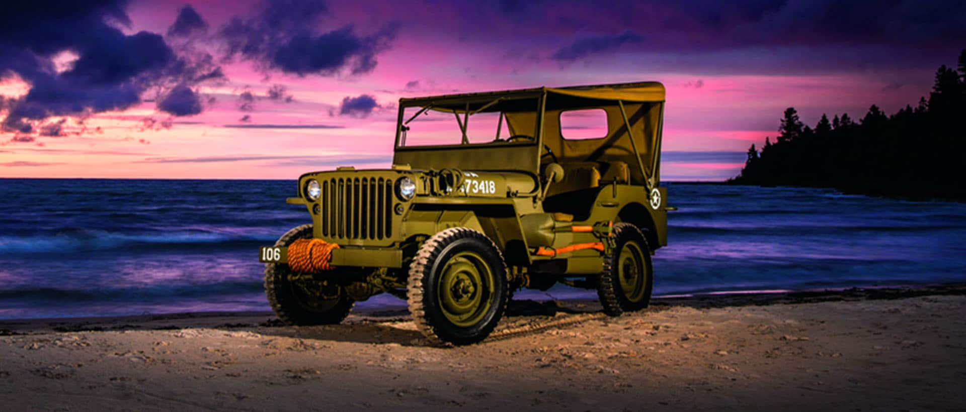 A World War 2 Jeep parked on a beach at sunset, with a pink and violet sky in the background.