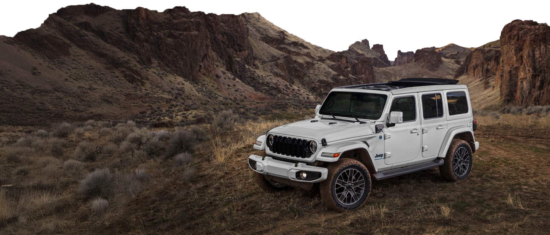 A white 2025 Jeep Wrangler Sahara 4xe parked on a clearing off-road among hills and outcroppings.