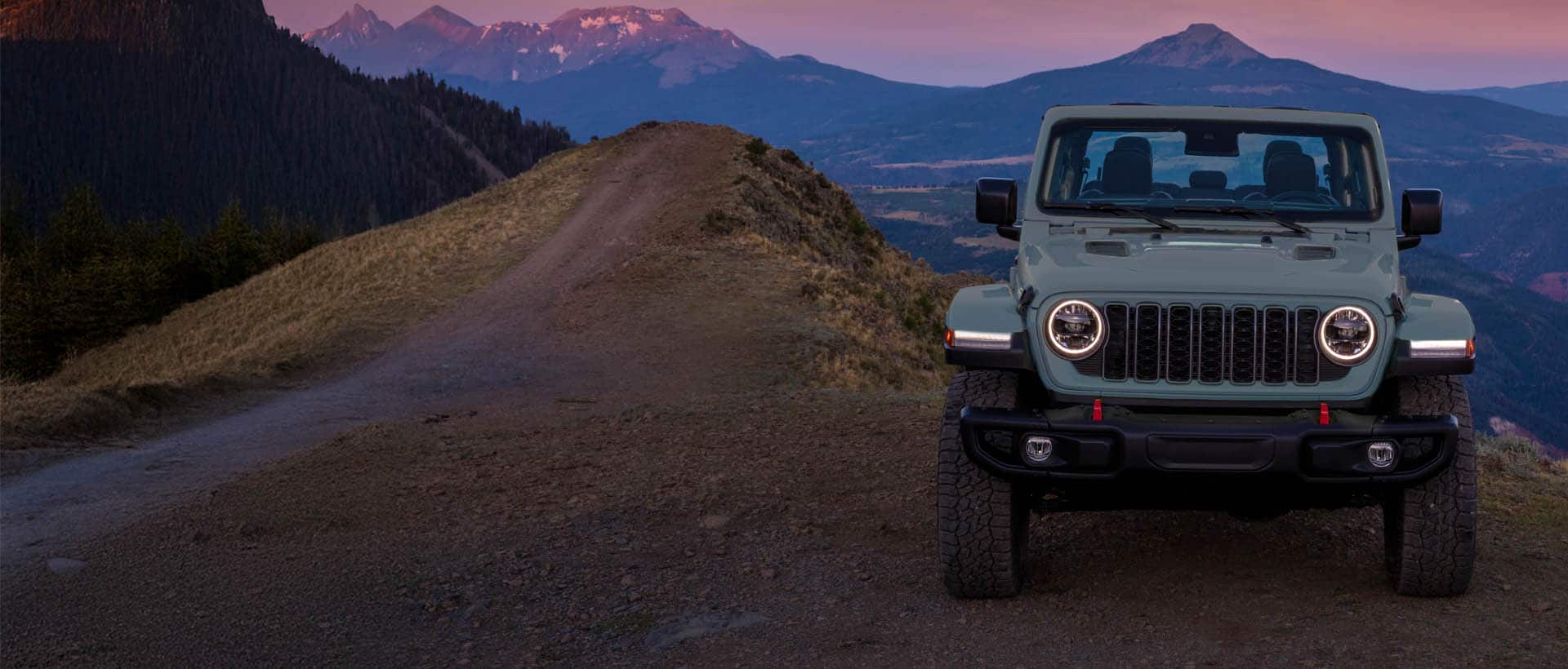 A blue gray 2025 Jeep Wrangler Rubicon X parked on a clearing in the mountains at sunset.