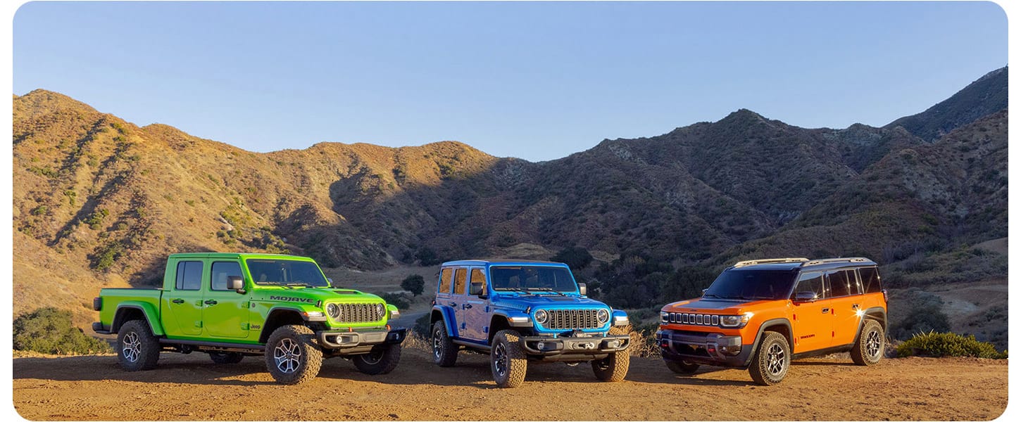 Three 2025 Jeep Brand vehicles on a clearing in the mountains. From left to right: A green Jeep Gladiator Mojave X, a blue Jeep Wrangler Rubicon X 4xe and an orange Jeep Recon preproduction model.