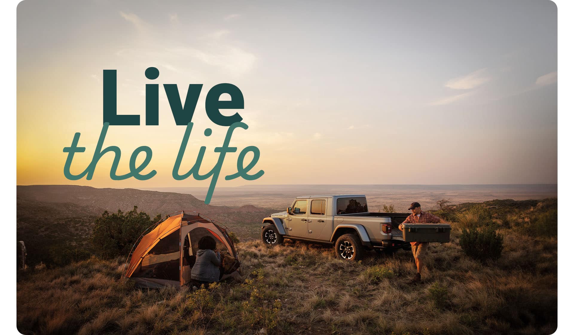 A 2025 Jeep Gladiator Rubicon X on a grassy clearing in the mountains with a man carrying a cooler and and a woman at a tent nearby.