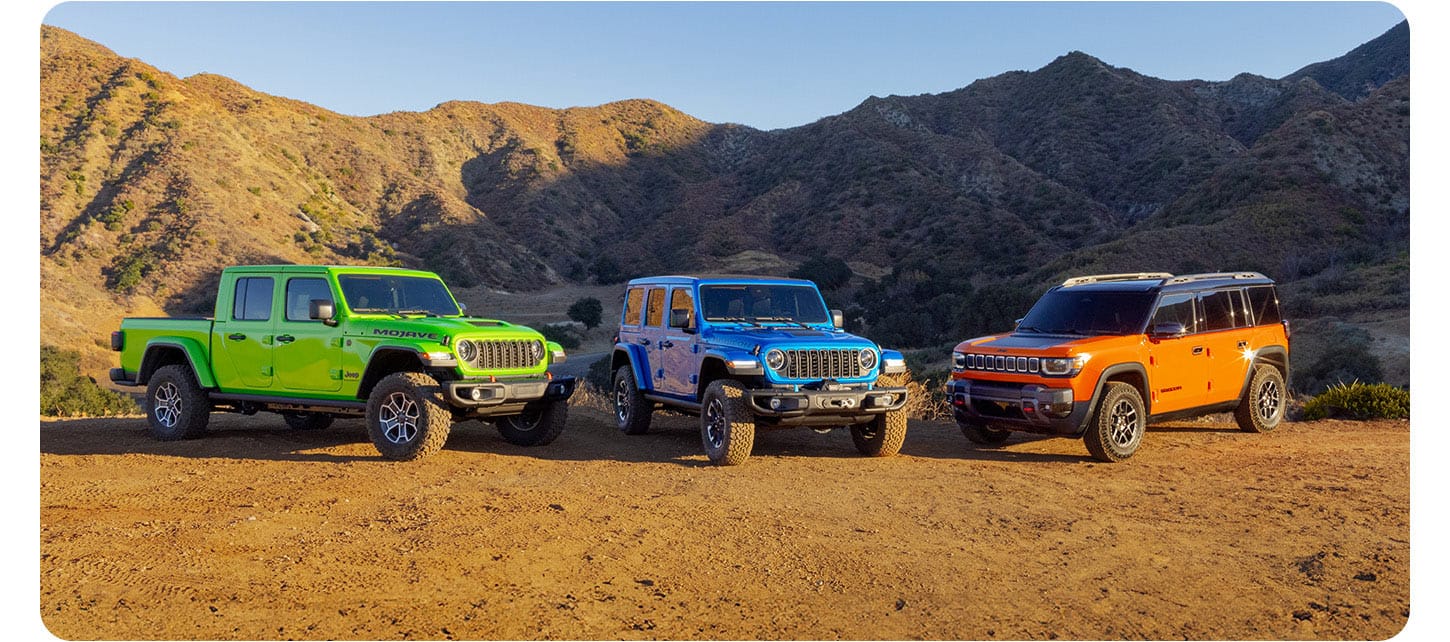 Three 2025 Jeep Brand vehicles on a clearing in the mountains. From left to right: A green Jeep Gladiator Mojave X, a blue Jeep Wrangler Rubicon X 4xe and an orange Jeep Recon preproduction model.