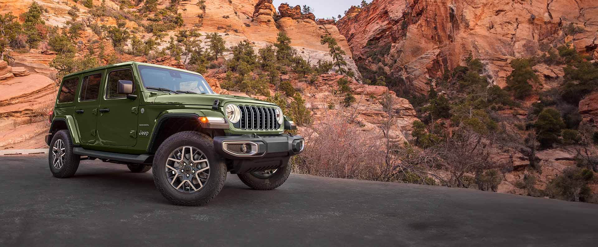 A green 2024 Jeep Wrangler Sahara parked on a paved clearing in the mountains.