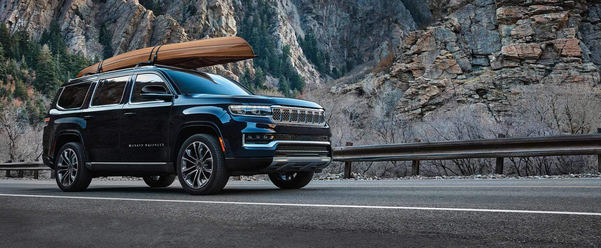 A black 2025 Jeep Grand Wagoneer Series III with a canoe strapped to its roof rack, parked on the shoulder of a mountain highway.