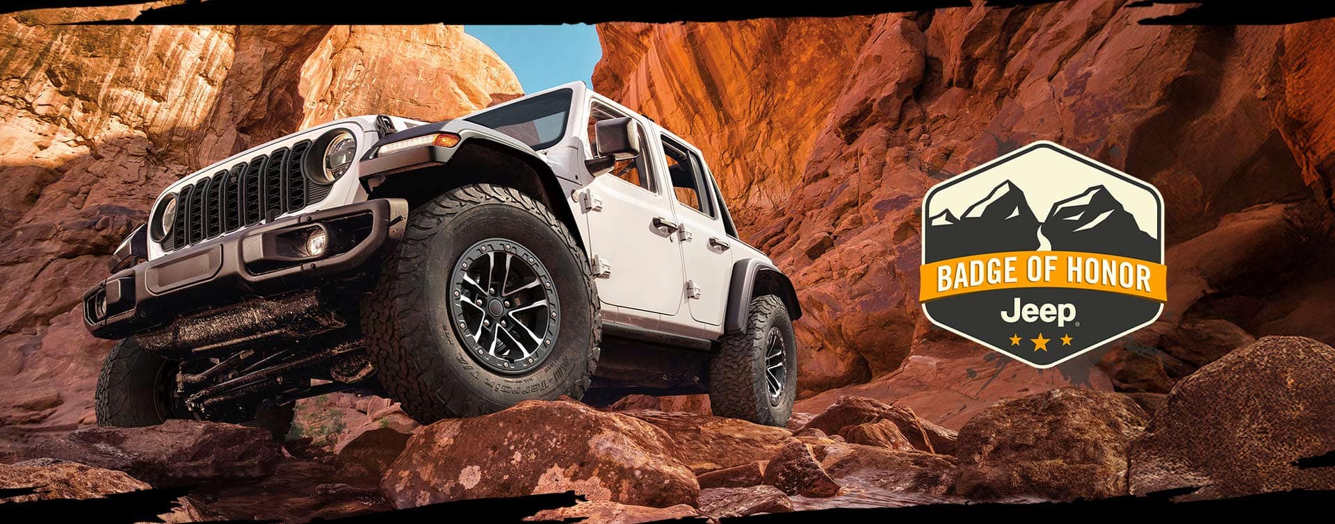 A 2026 Jeep Wrangler Rubicon, seen from below as it surmounts a rock in a canyon. Jeep Badge of Honor.