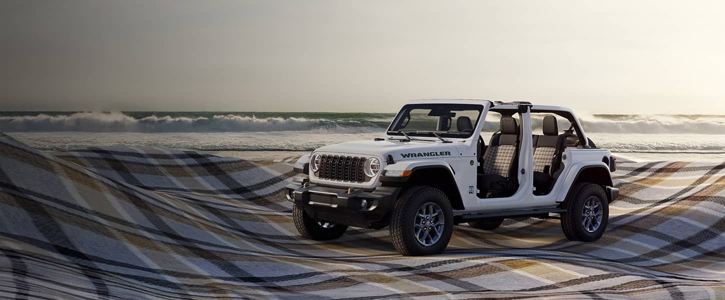 A driver-side front angle of a white 2026 Jeep Wrangler 85th Anniversary Edition with its doors and top removed, revealing plaid inserts on the front and rear seats. It is parked beside the ocean on a massive billowing plaid blanket.
