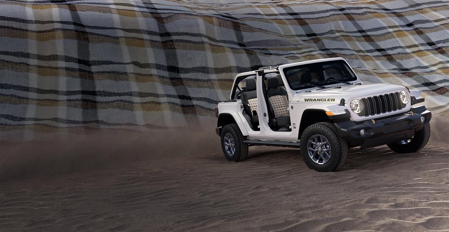 A passenger-side front angle of a white 2026 Jeep Wrangler 85th Anniversary Edition with its doors and top removed, revealing plaid inserts on the front and rear seats. It is parked on sand, with a massive plaid blanket as a backdrop.