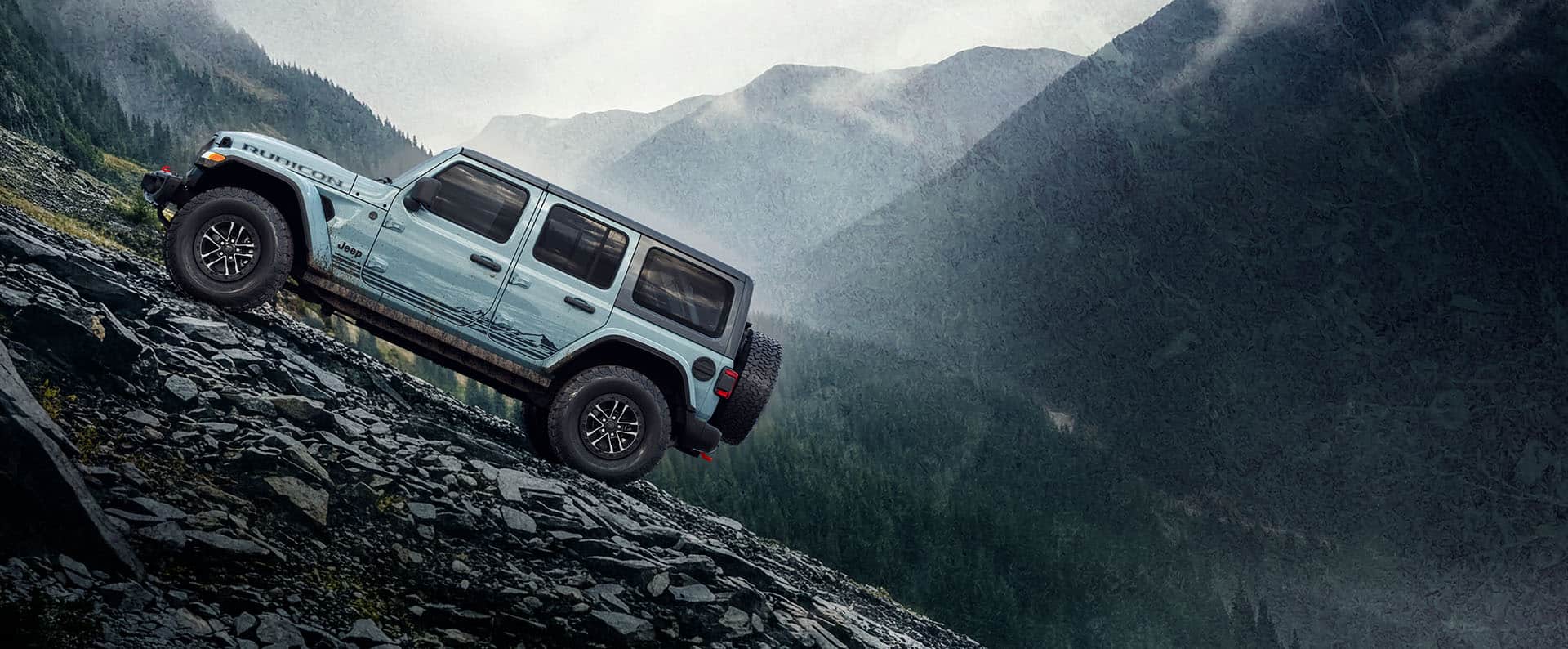 A driver-side profile of a blue-green 2026 Jeep Wrangler Rubicon Rockslide crawling up a steep, rocky trail off-road with misty mountains in the background.