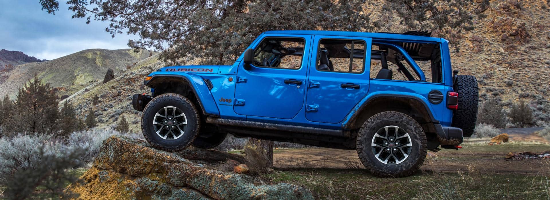 A driver-side profile of a blue 2026 Jeep Wrangler Rubicon X four-door, parked off-road, with its front tires elevated on a raised rock formation.